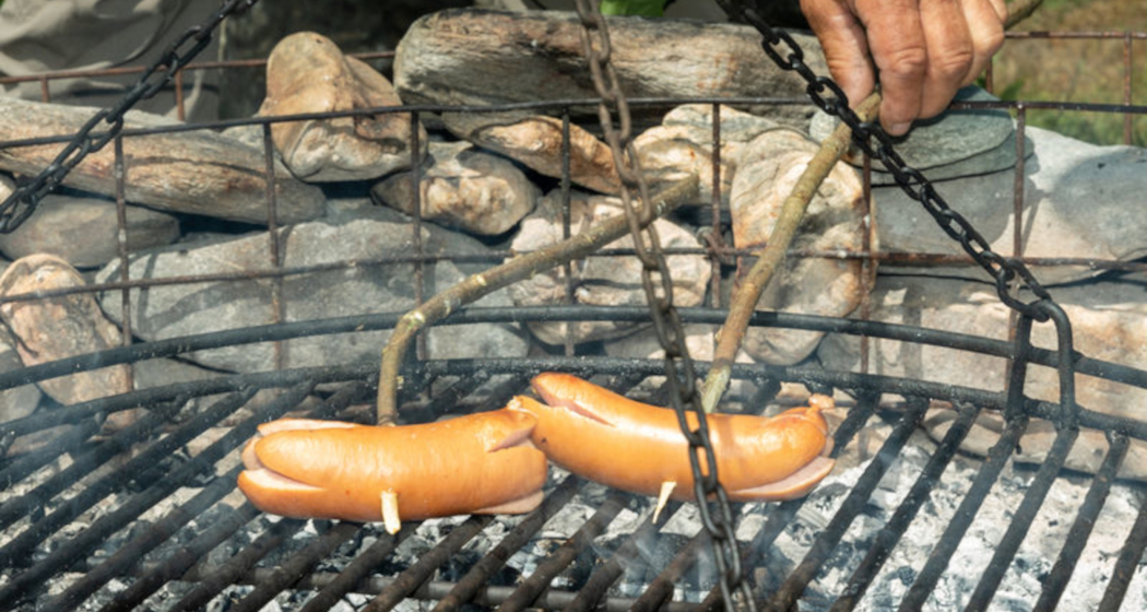 Das Foto zeigt eine Feuerstelle und zwei Cervelats die am braten sind Das Foto zeigt eine Feuerstelle und zwei Cervelats die am braten sind